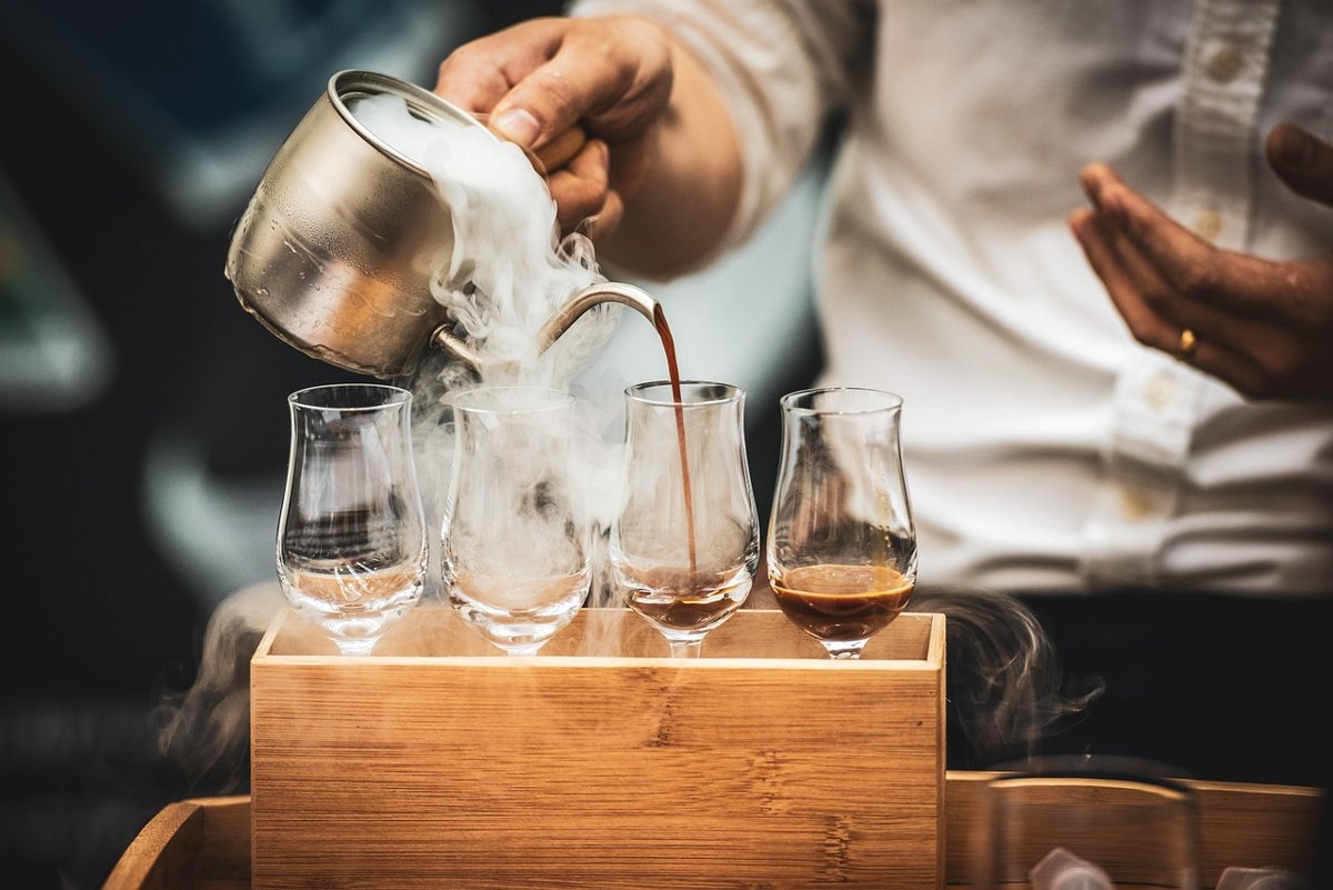 Barista pouring hot water from a kettle into a brewer on a wooden coffee bar, with glasses lined up in front
