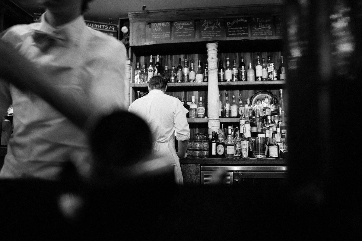 Black and white photo of a bartender working in front of shelves of bottles in a dim cocktail bar