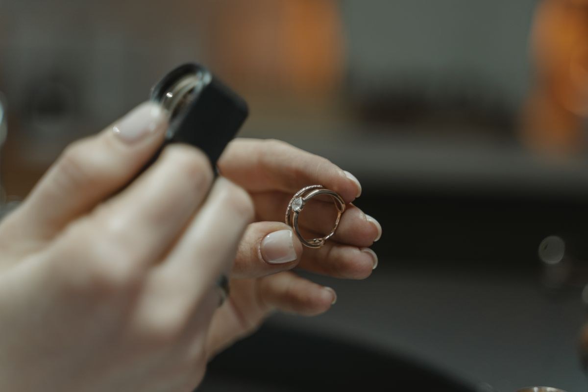 Fingers holding a slim gold ring with a small clear centre stone next to a black jeweller's loupe