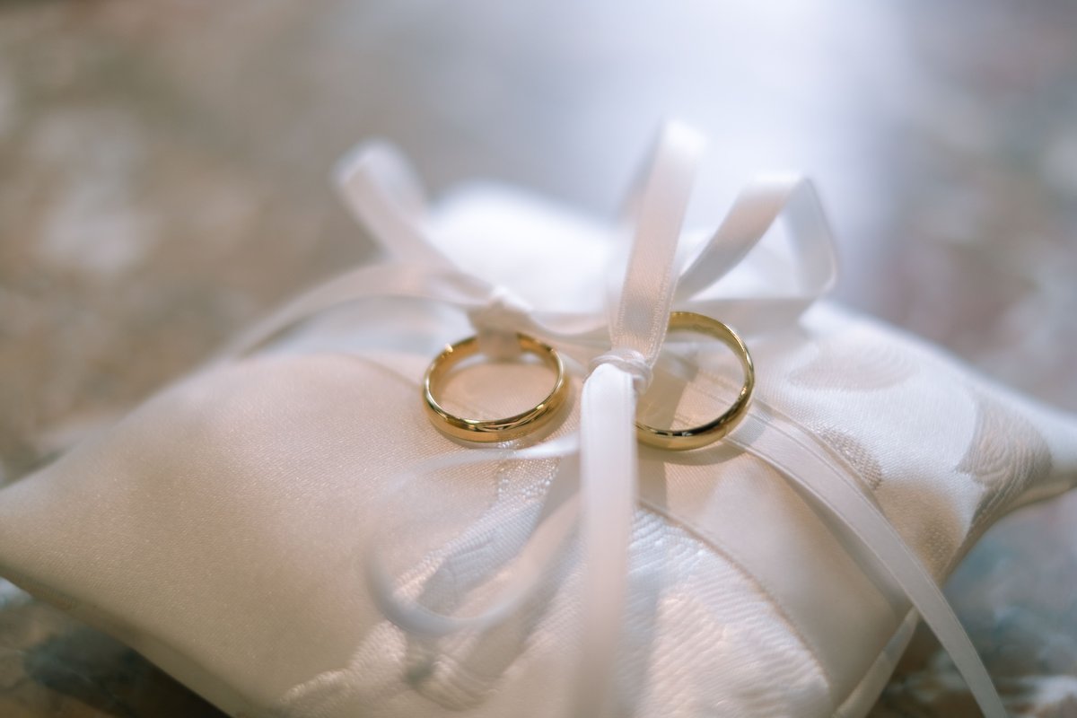 Two plain polished gold wedding bands tied with white satin ribbon on a cream ring pillow