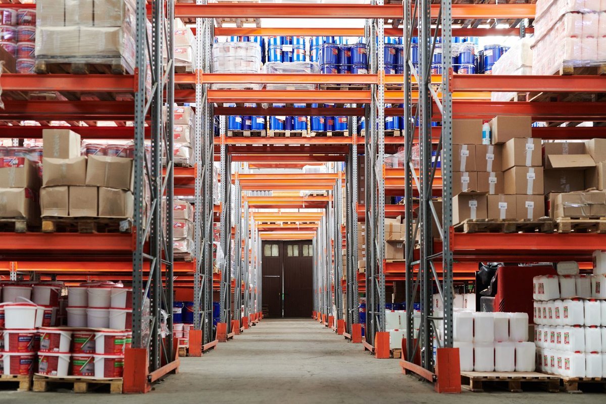 Long warehouse aisle lined with orange pallet racking stacked with boxes and drums
